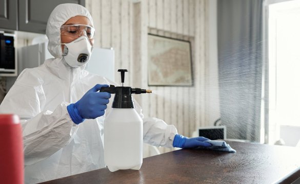 Professional cleaner in protective suit disinfecting a surface with a spray bottle during deep cleaning.